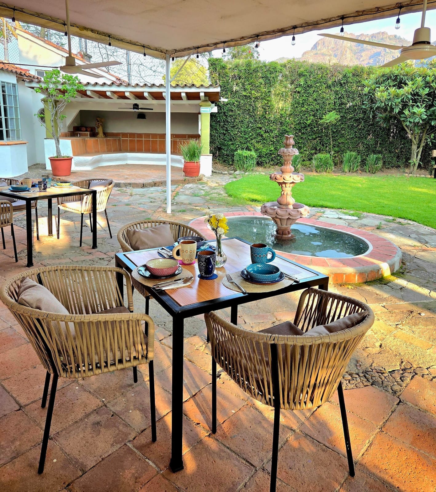 Outdoor patio dining area with wicker chairs and wooden tables set for a meal under a canopy and string lights, with a stone fountain and red-brick edging nearby.